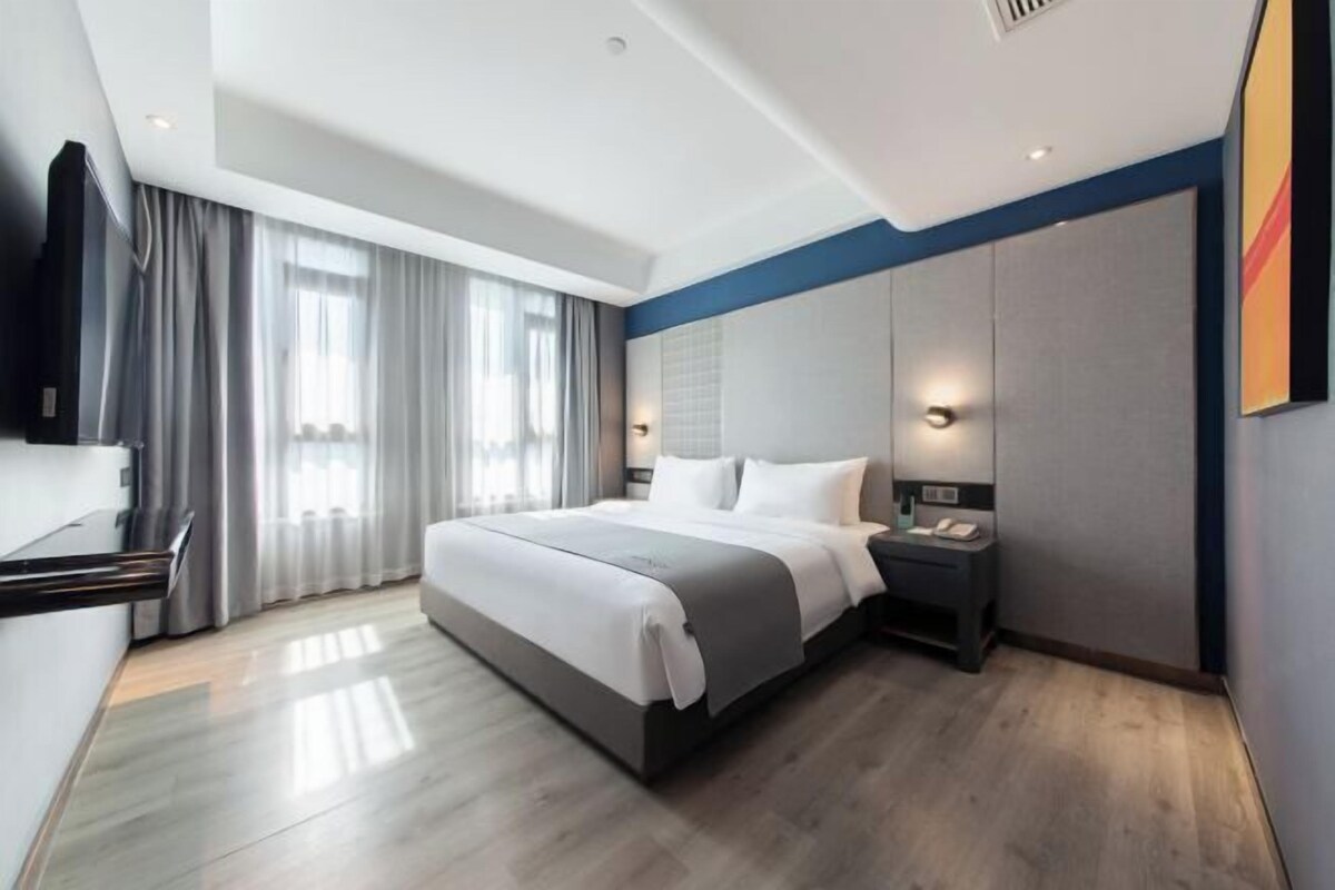 Une chambre d’hôtel moderne dotée d’un grand lit, de tables de chevet, d’un téléviseur à écran plat et d’une grande fenêtre avec des rideaux.