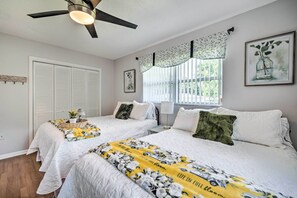 Ein Hotelzimmer mit zwei Betten, einem Deckenventilator, einem Fenster mit Jalousien, einem gerahmten Bild an der Wand und einem Korb mit einer Pflanze auf dem Nachttisch.