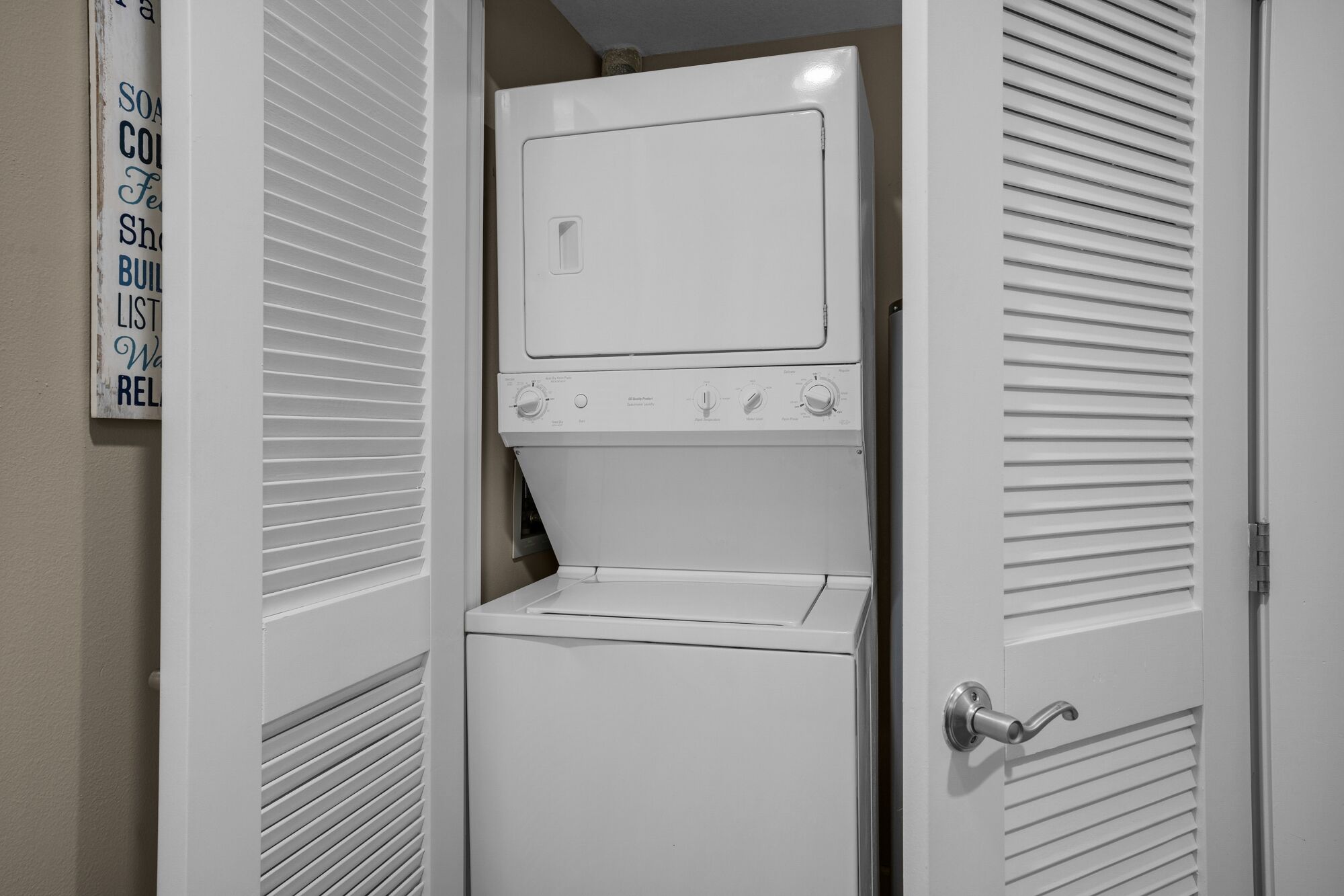 A stacked washer and dryer unit in a laundry room with a door handle and a sign on the wall.