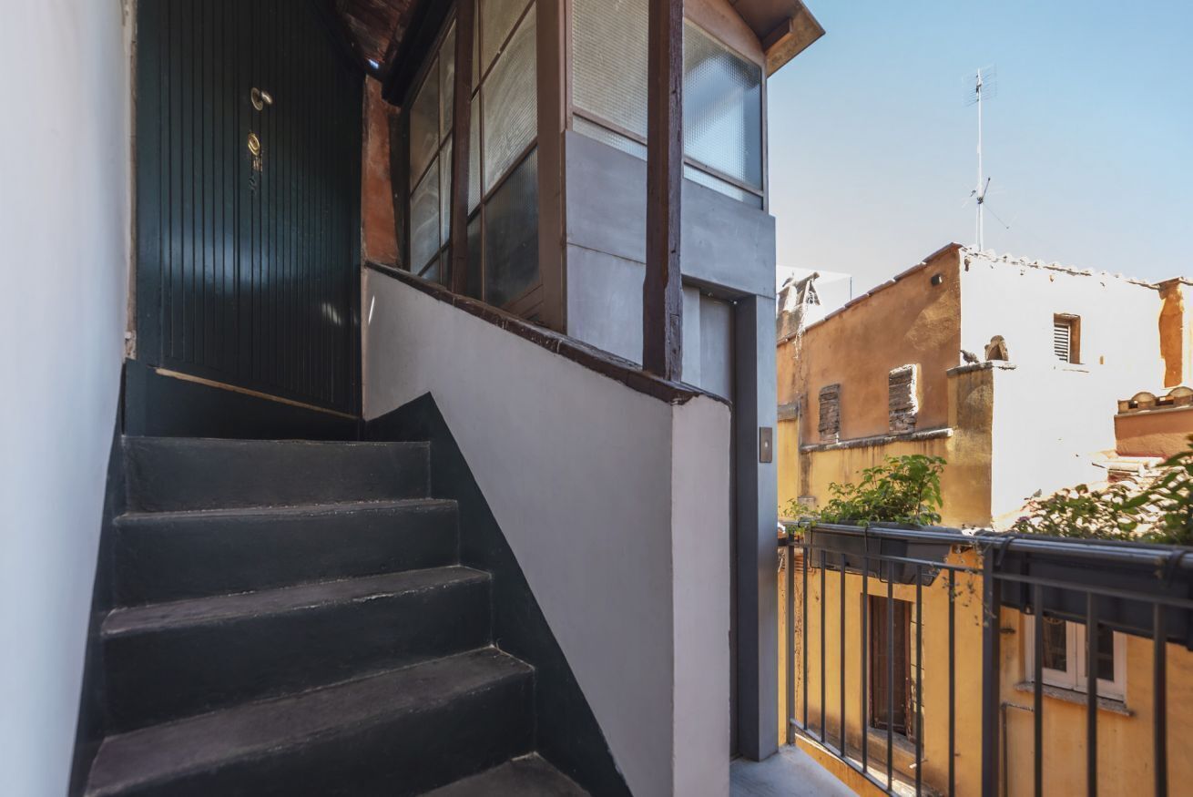 A staircase leading to a dark wooden door, adjacent to a white wall and a balcony with a black railing.