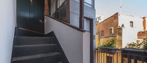 A staircase leading to a dark wooden door, adjacent to a white wall and a balcony with a black railing.