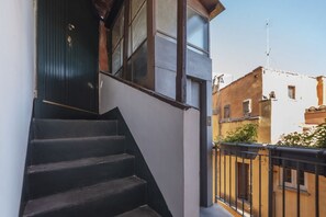 Eine Treppe führt zu einer dunklen Holztür, neben einer weißen Wand und einem Balkon mit schwarzer Geländer.