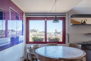 A dining area with a round table and chairs, large windows with a view of buildings, and a pendant light.