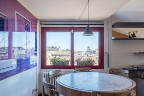 A dining area with a round table and chairs, large windows with a view of buildings, and a pendant light.