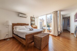 A bedroom with a wooden bed, a wicker bench, a floor lamp, and a view of the city through large windows.