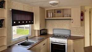 A compact kitchen with a gas stove, a built-in oven, a window with a curtain, and a fire extinguisher mounted on the wall.