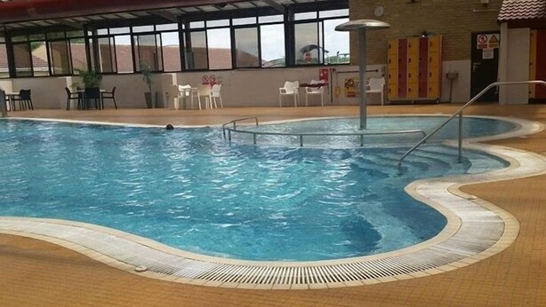 An indoor swimming pool with a tiled floor and a poolside area with chairs and tables.