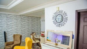 A living room with a stone wall, a clock, a yellow armchair, a brown armchair, a dining table with chairs, and a cabinet with a mirror. - Beautiful 4-Bedroom House Located in Abuja (Abuja)