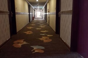 Un pasillo de hotel con alfombra estampada con motivos de hojas, puertas a ambos lados y una cortina púrpura a la derecha.
