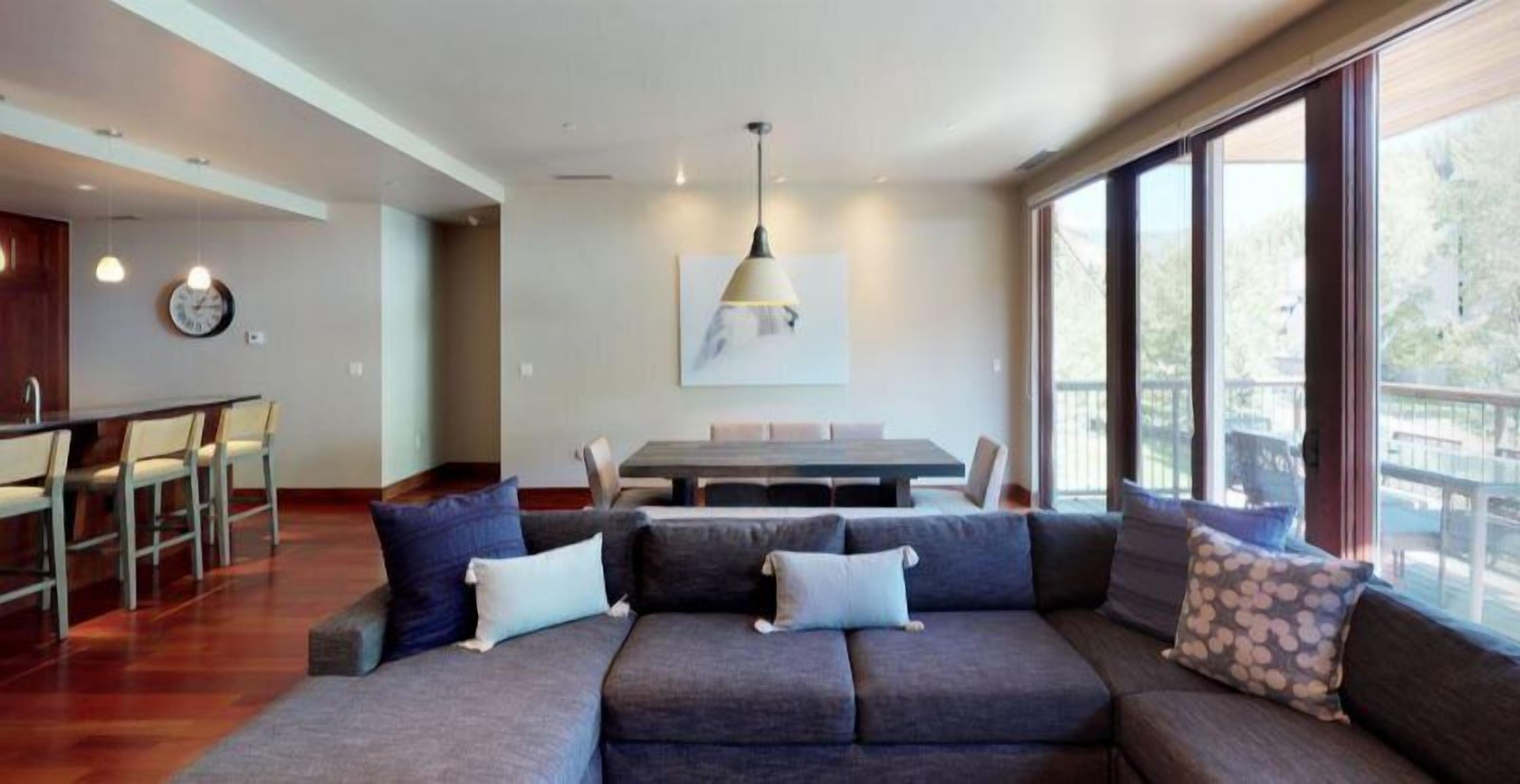 A modern living room with a sectional sofa, dining table, and chairs. There is a large window providing a view of the outdoors.