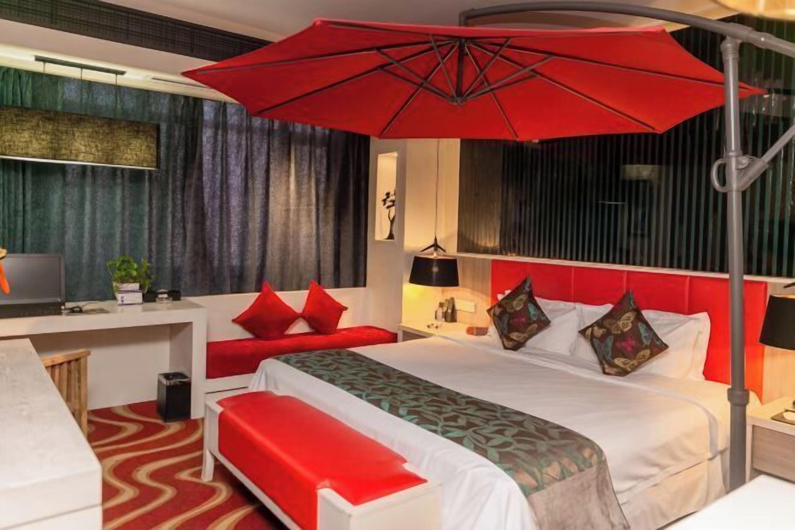 A hotel room with a bed, a desk, a red umbrella, and a red bench.