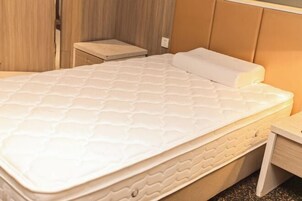 Una cama bien tendida con un colchón y almohada blancos, una mesita de noche de madera a la derecha y un cabecero de madera.