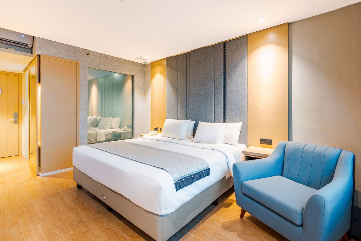 A modern hotel room with a large bed, a blue armchair, and a glass partition wall.