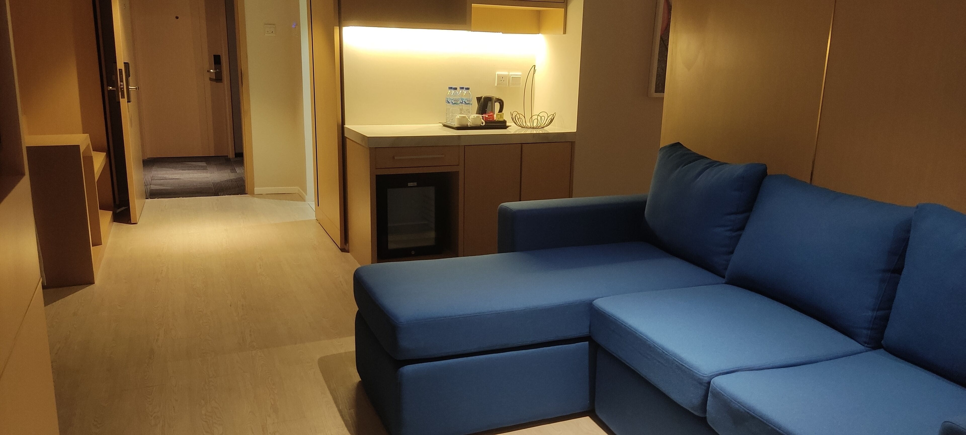 Une chambre d’hôtel moderne avec un canapé d’angle bleu, une petite kitchenette et un réfrigérateur compact.