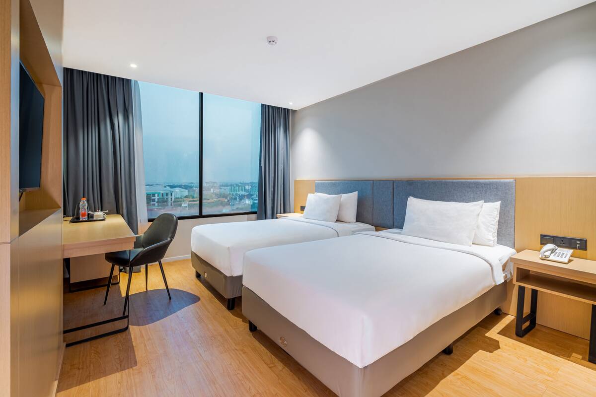 A hotel room with two beds, a desk, a chair, and a large window offering a city view.