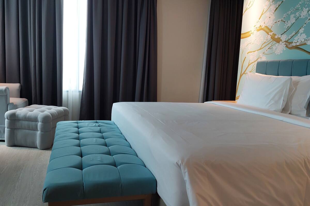 A hotel room with a bed, a chair, an ottoman, and a large window with curtains.