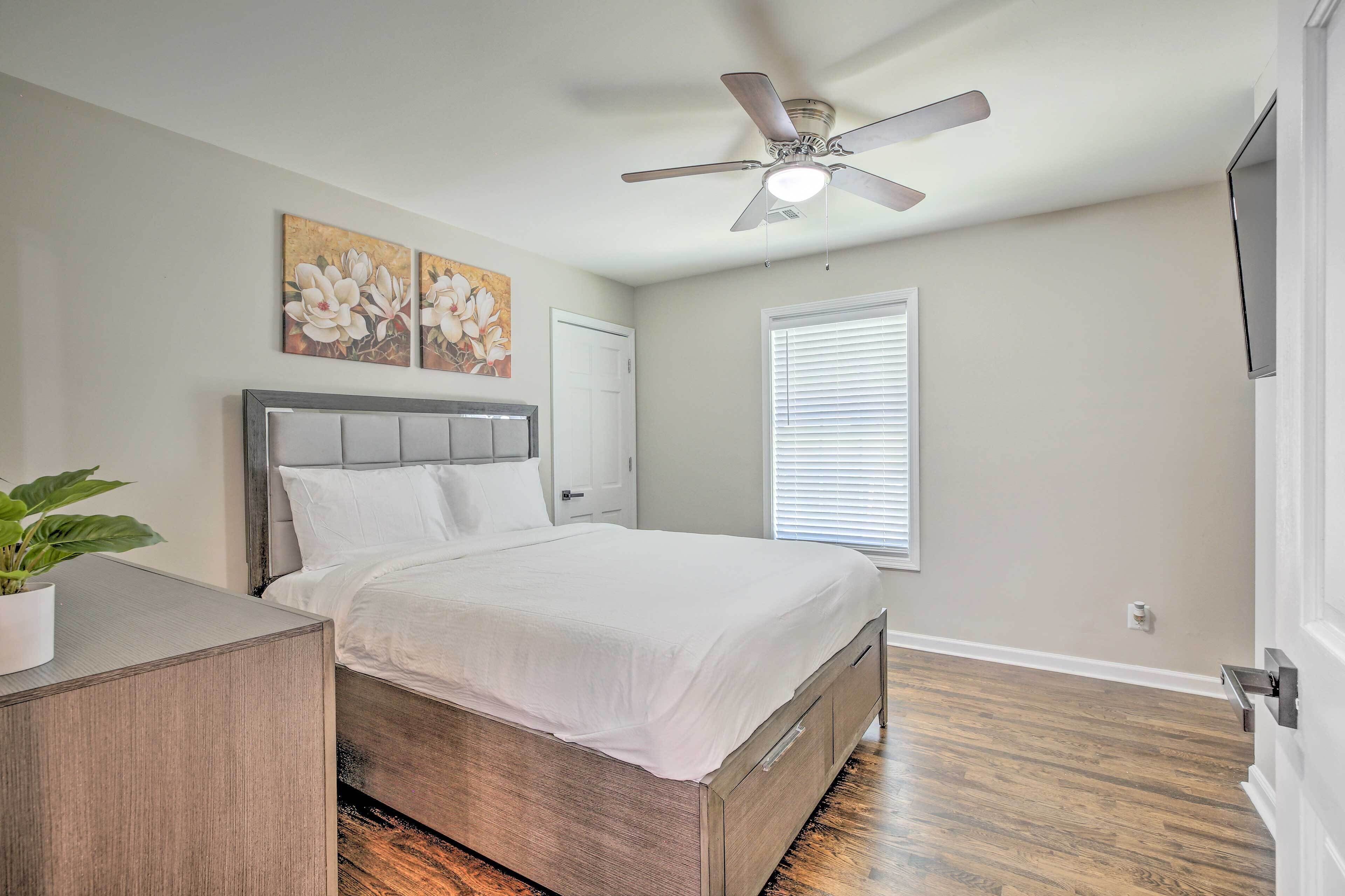 A bedroom with a bed, a ceiling fan, a window with blinds, and a wall-mounted TV.