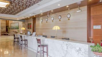 A hotel reception area with a marble counter, several chairs, a clock, and a potted plant.