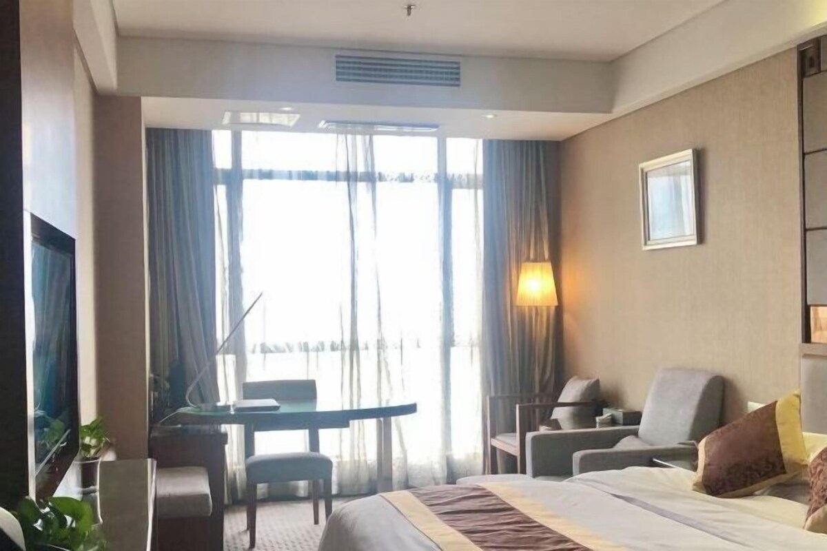 A hotel room with a bed, a desk, two chairs, a TV, and a window with curtains.