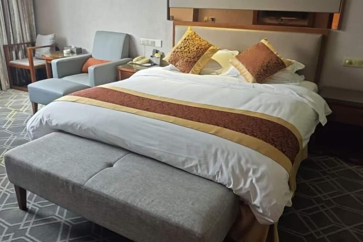 A hotel room with a bed, a grey armchair, a small table with a lamp, and a nightstand with a phone.