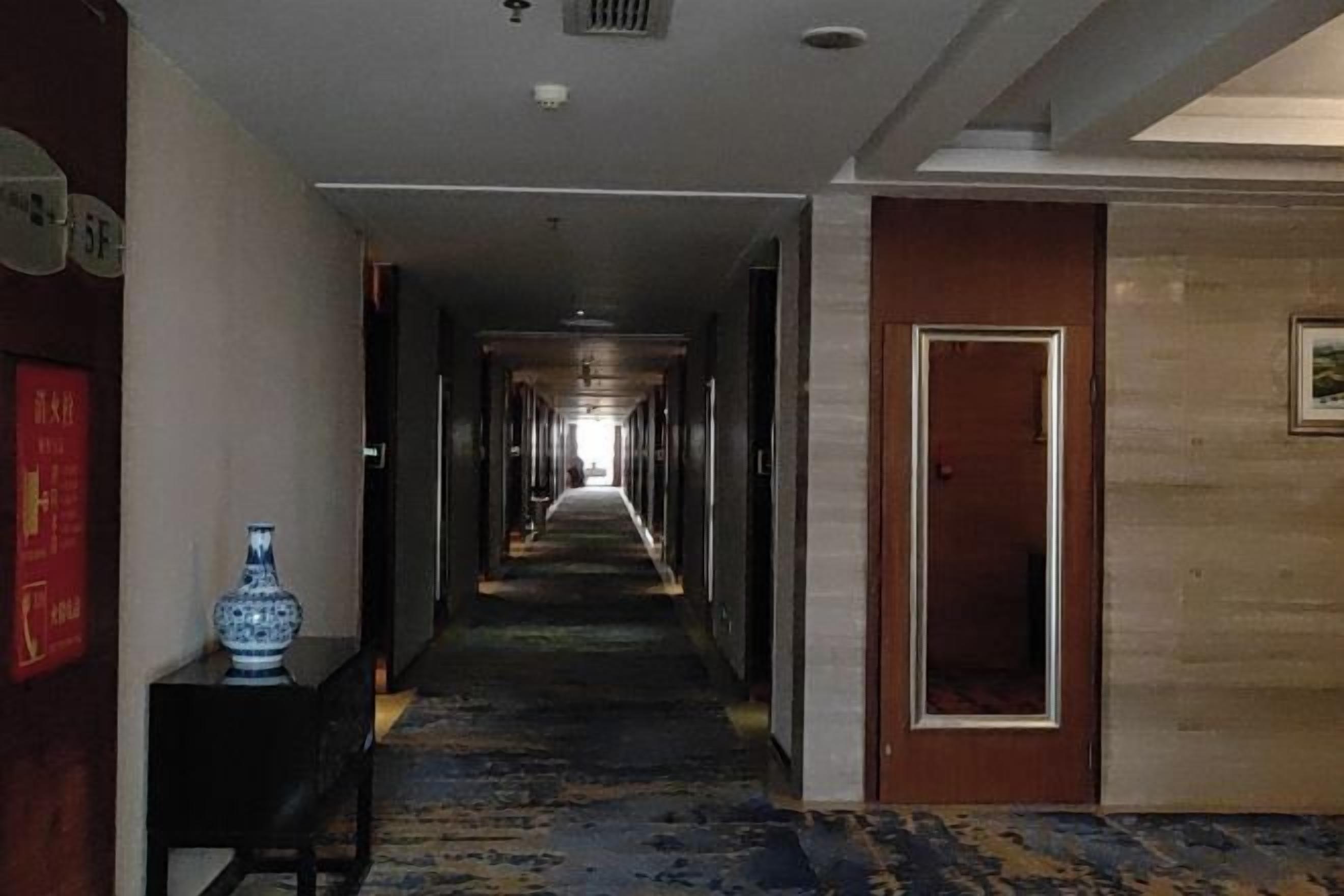 A hotel corridor with a carpeted floor, a vase on a table, and a door at the end.
