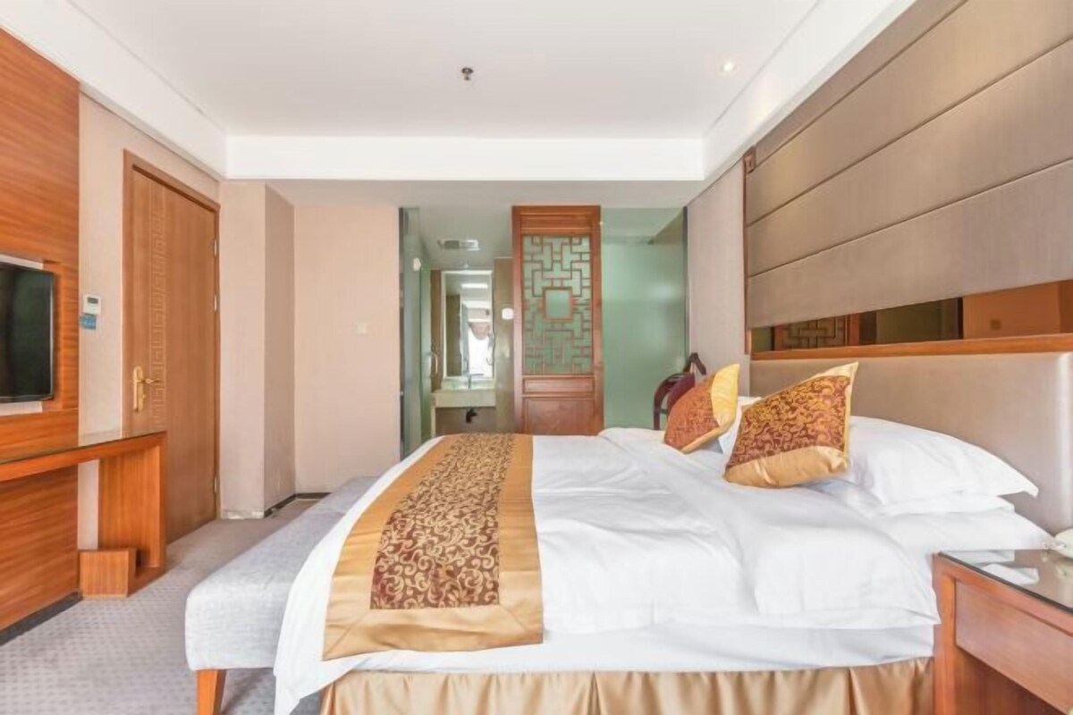 A hotel room with a large bed, a wooden headboard, a TV on the left, and a wooden wardrobe.