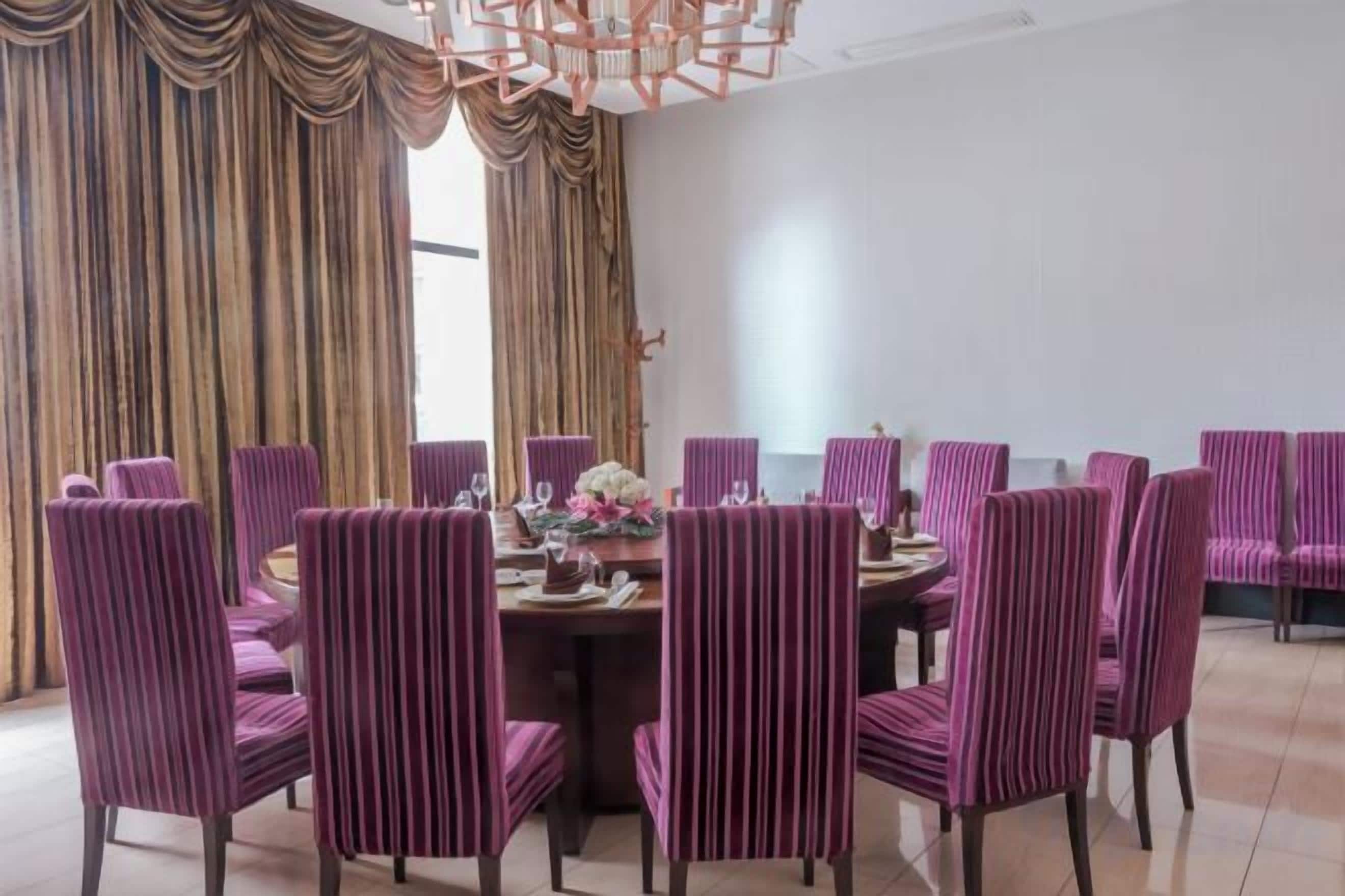 Un comedor con una mesa redonda rodeada de sillas tapizadas en color morado, una araña de cristal y cortinas.