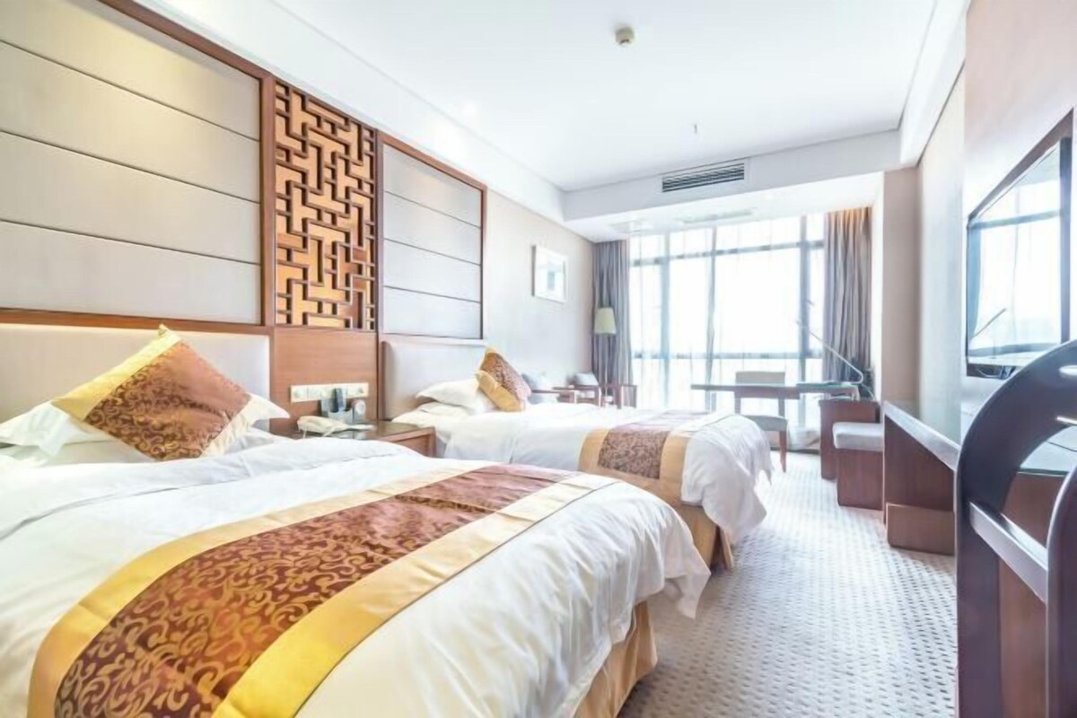 A hotel room with two beds, a TV, a desk, and a large window.