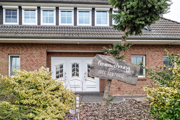 A brick house with a white door and windows, a signboard with contact information, and surrounding greenery.