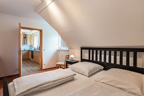 A bedroom with two beds, a nightstand, a chair, and a view of a kitchen and bathroom through an open door.