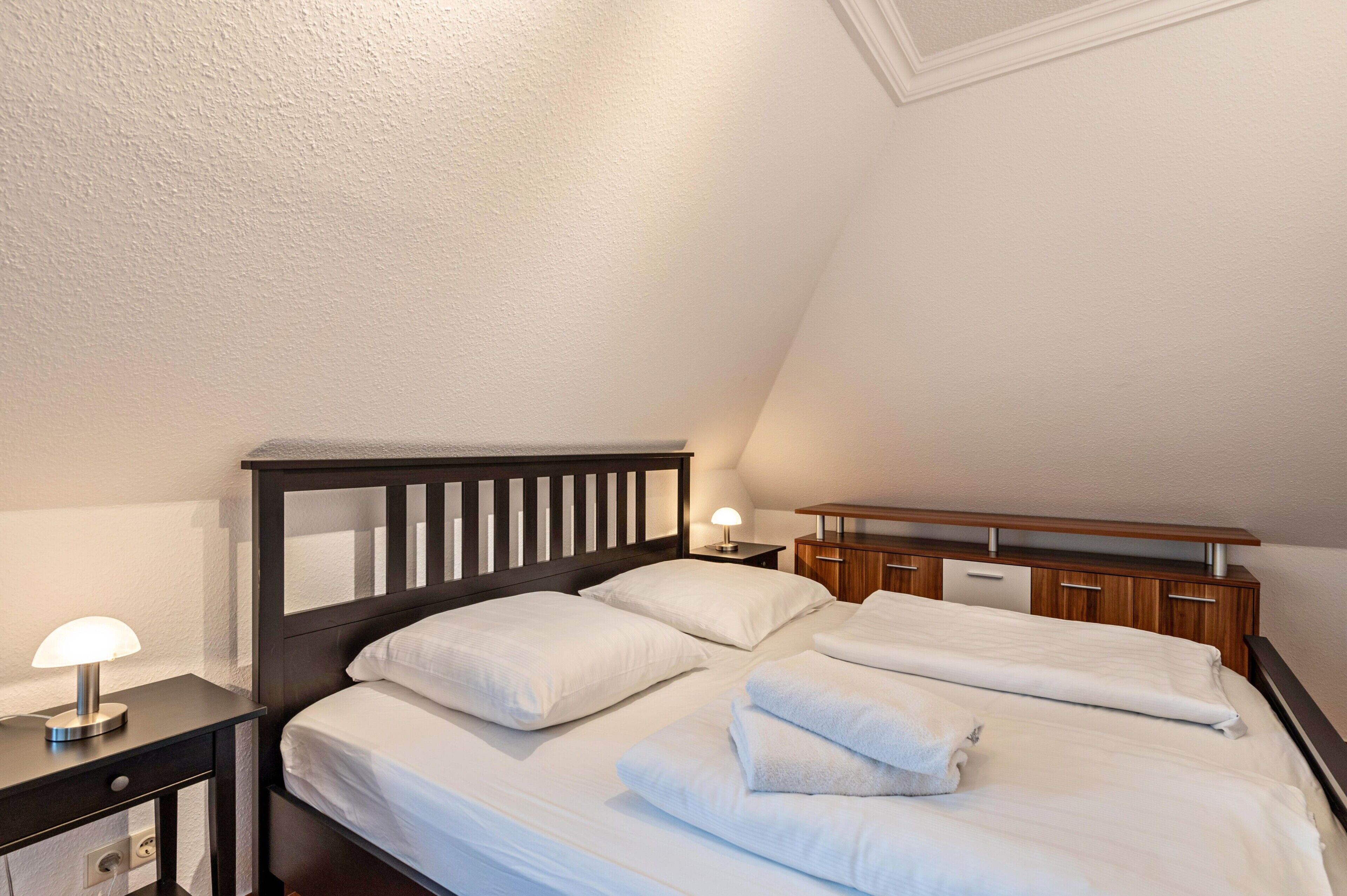 Een net opgemaakt bed met witte lakens, een houten hoofdbord en een nachtkastje met een lamp.