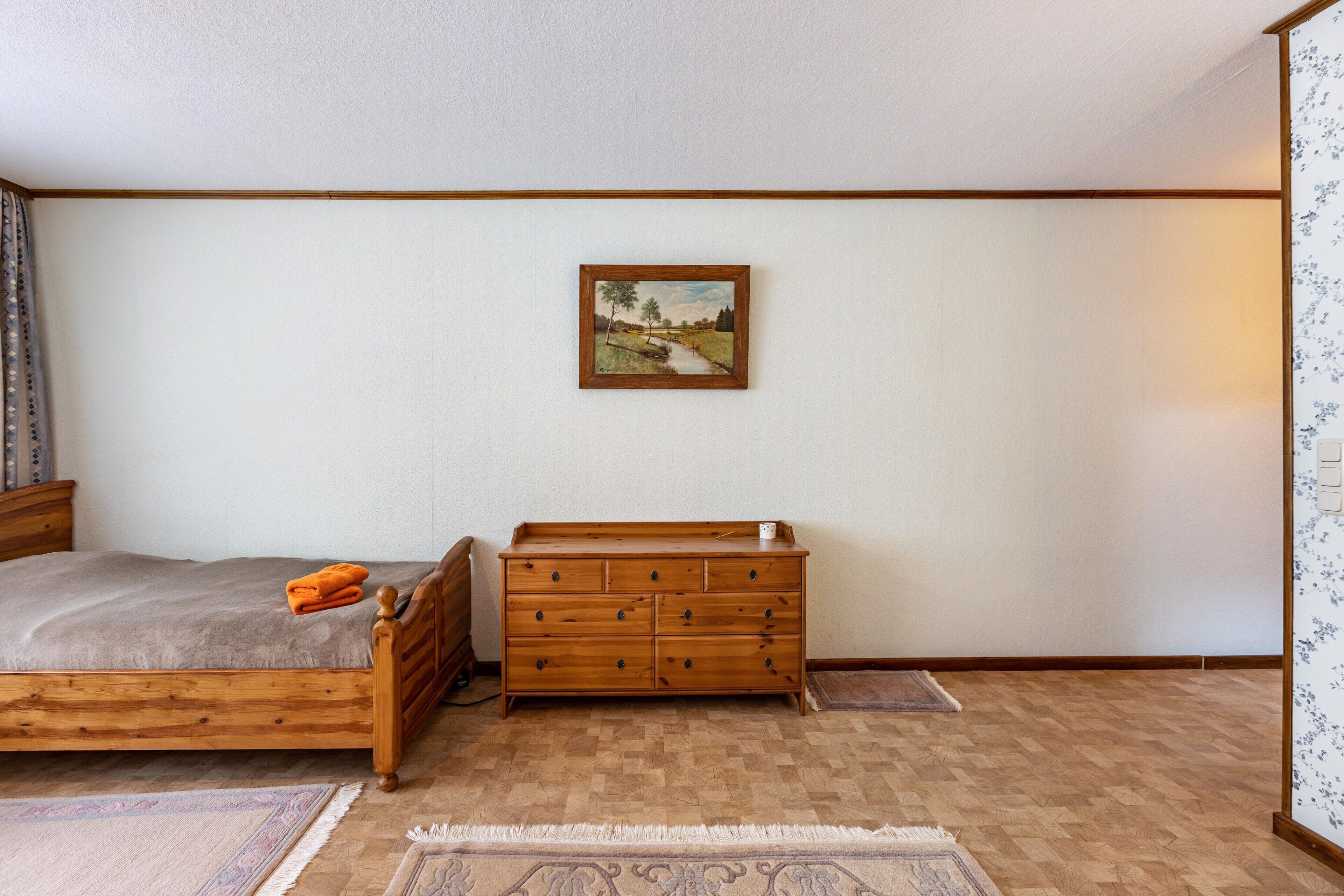 Une chambre à coucher avec un lit en bois, une commode, un tableau accroché au mur et un tapis à motifs sur le sol.