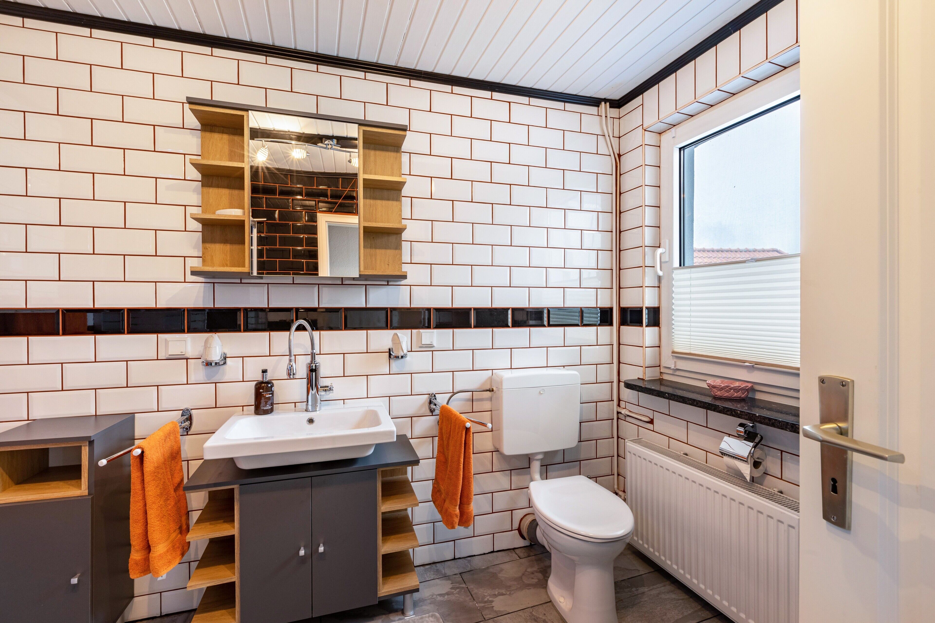 Ein modernes Badezimmer mit weiß gefliesten Wänden, einem Waschbecken, einer Toilette und einem Fenster.