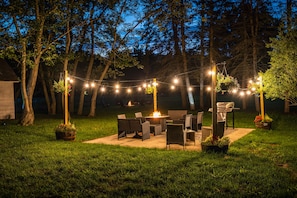 Un patio al aire libre con mobiliario de mimbre y luces colgantes, rodeado de árboles.