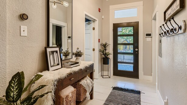 A hallway with a white textured wall, a dark door, a mirror, a light fixture, and a plant.