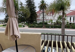 Un balcón con una sombrilla color beige y una mesa redonda, con vista a una calle con palmeras y un edificio grande con techo rojo.