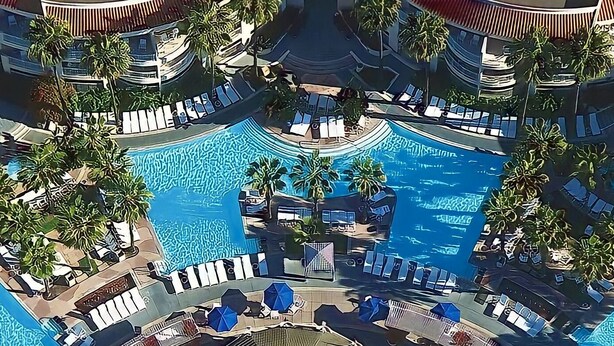 An aerial view of a resort with a central swimming pool surrounded by lounge areas and palm trees.