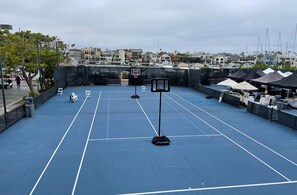 Una cancha de baloncesto al aire libre con aro, rodeada por una valla, con vistas a un puerto deportivo con barcos y casas.