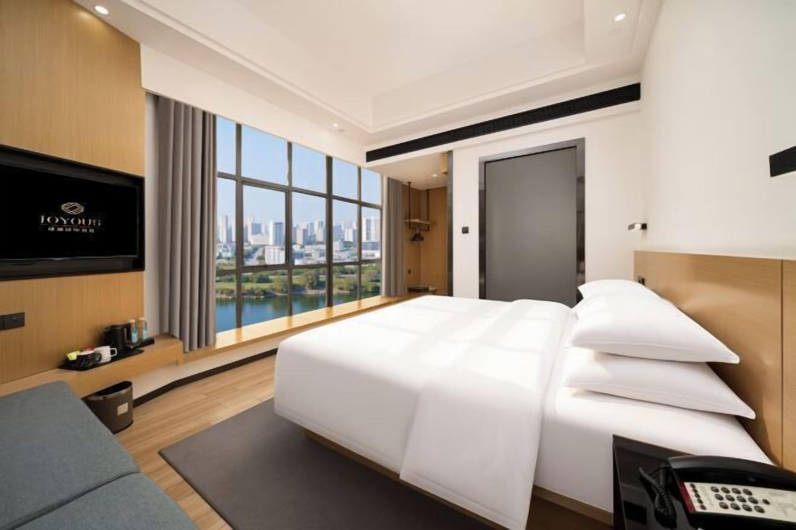 A modern hotel room with a large bed, a TV mounted on the wall, a city view, and a cityscape visible through the window.