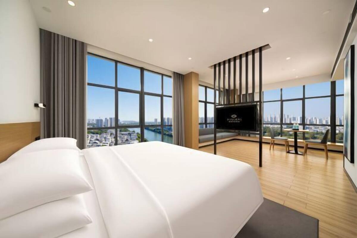 A modern hotel room with a large bed, a TV, a balcony with a city view, and a large window.