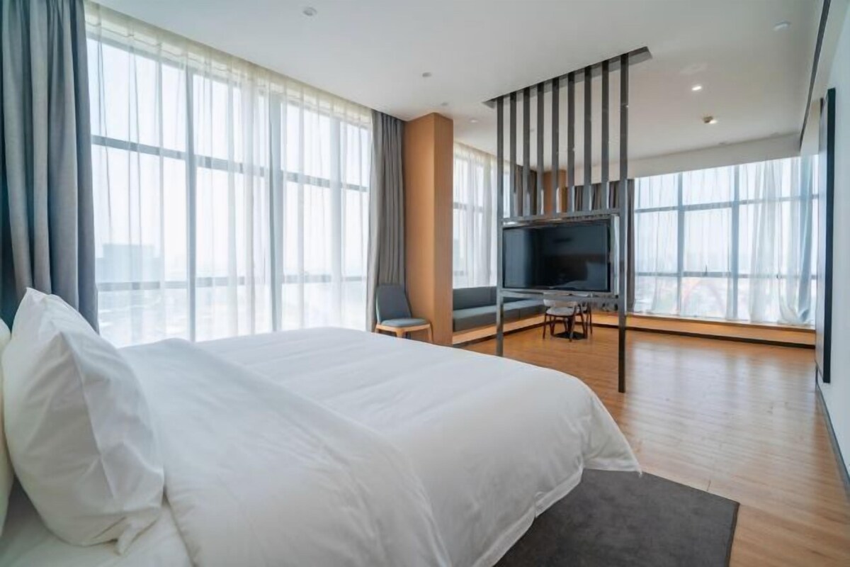 Una habitación de hotel moderna con una cama grande, un escritorio, un televisor y ventanas amplias.