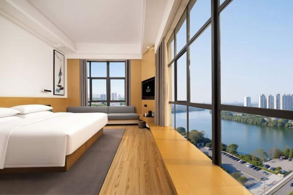 A modern hotel room with a large bed, a seating area, a TV, and a view of a cityscape and water.
