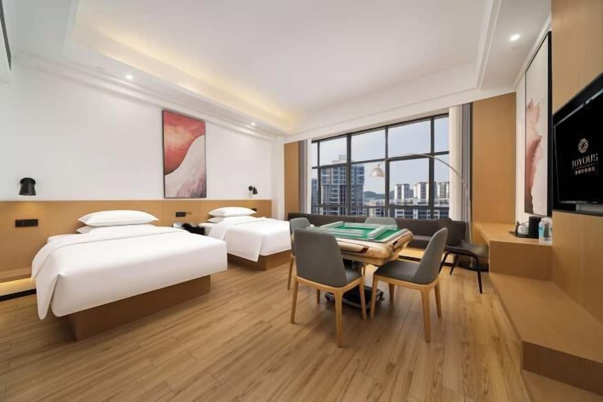 A hotel room with two beds, a desk with chairs, a large window, and a television.