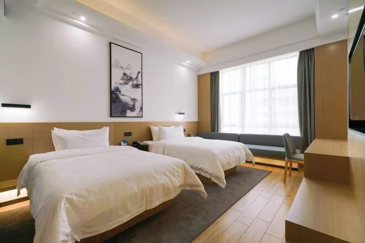 A hotel room with two beds, a desk, a chair, a large window, and a painting on the wall.