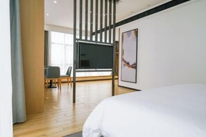 Moderni hotellihuone, jossa on suuri sänky, seinälle asennettu taulutelevisio, oleskelualue tuoleineen ja suuri ikkuna, josta tulee runsaasti luonnonvaloa.
