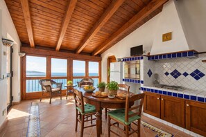 A kitchen with wooden beams, a dining area with chairs, a table with food, and a view of the sea. - Casa Lella, Furore, Italy (Furore)