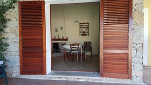 An open wooden door leading to a dining area with a table and chairs, a hanging light fixture, and a wall-mounted shelf with decorative items. - House / Villa - Villasimius (Villasimius)