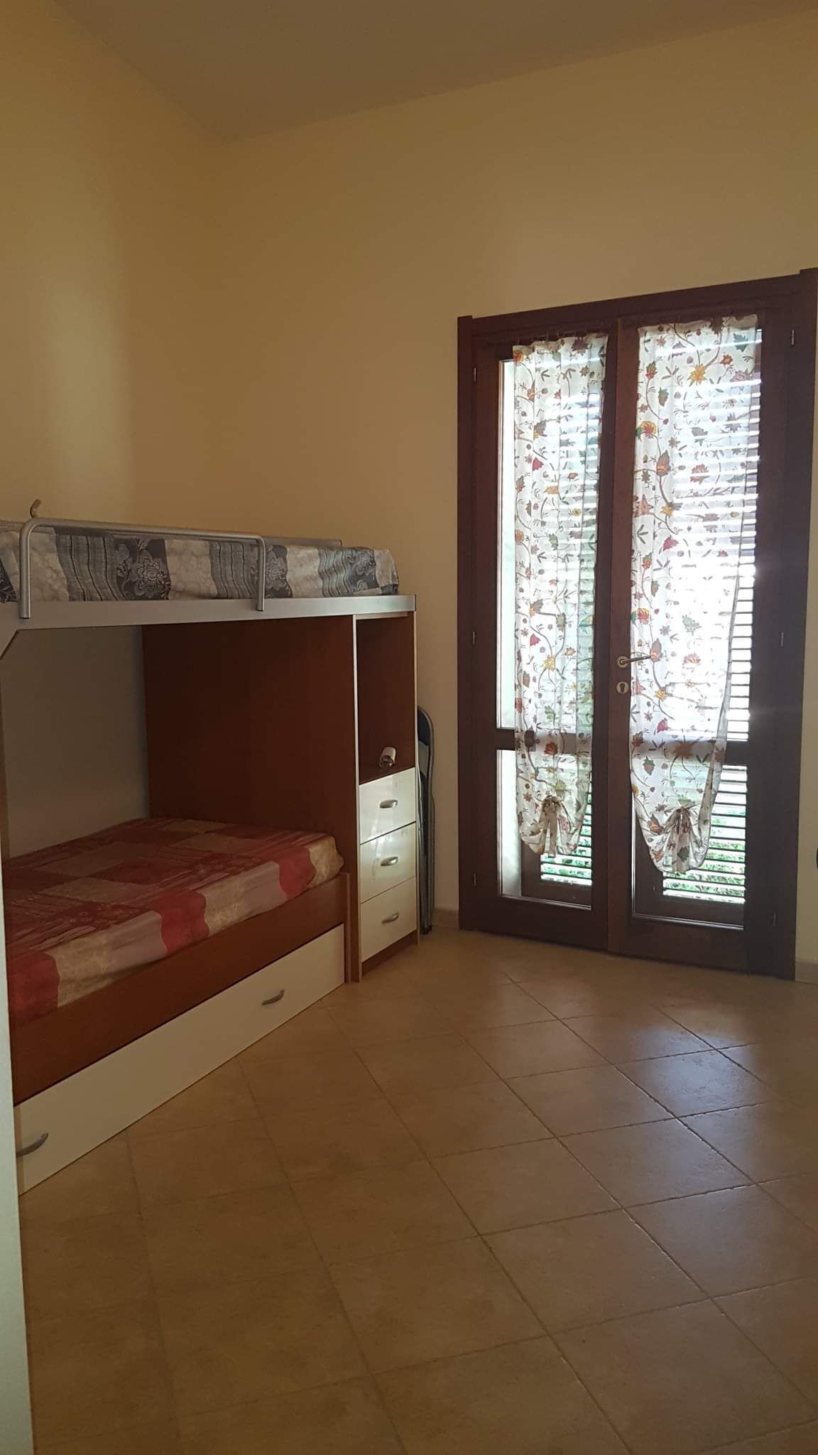 Una stanza con un letto a castello, una piccola scrivania e una porta scorrevole in vetro con tende floreali.