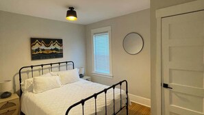 A bedroom with a bed, a bedside table, a lamp, a window with blinds, a wall-mounted mirror, and a door. - Trendy and Stylish 3BR Uptown Unit (Minneapolis)