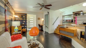 Un salon moderne avec un mur d’accent en bois, un ventilateur de plafond, un coin repas avec une table et des chaises, et un coin cuisine avec un comptoir et des étagères.